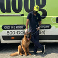 Load image into Gallery viewer, A Doog groomer posing proudly with a German Shepherd outside the mobile grooming van, showcasing a happy pet after a successful grooming session in a vibrant outdoor setting.